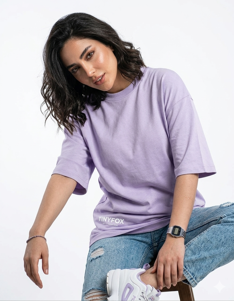 Woman in lavender oversized t-shirt with Tinyfox logo, ripped jeans, and white sneakers sitting on a stool against white background