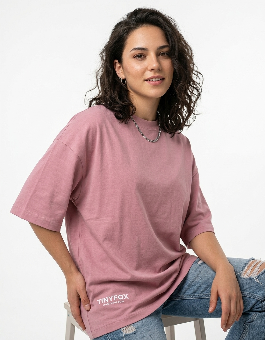 Woman in oversized pink T-shirt with Tinyfox logo and ripped jeans, sitting on a stool